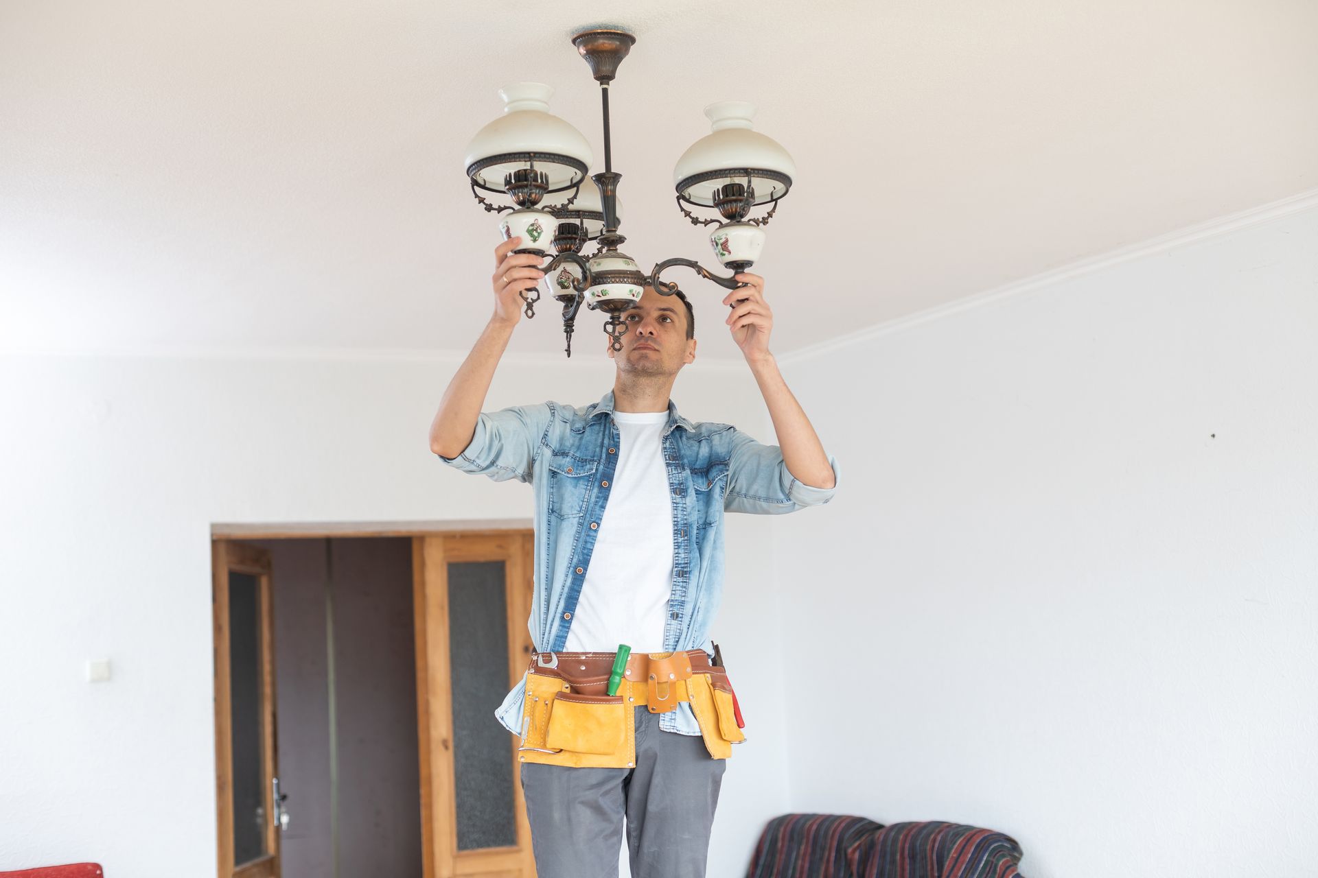 Man on a step stool installs a chandelier in a white room. He wears a tool belt and denim shirt.