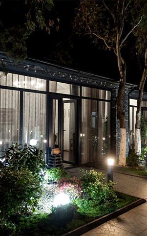 Night exterior view of a building with large windows, landscaping, and pathway lights.