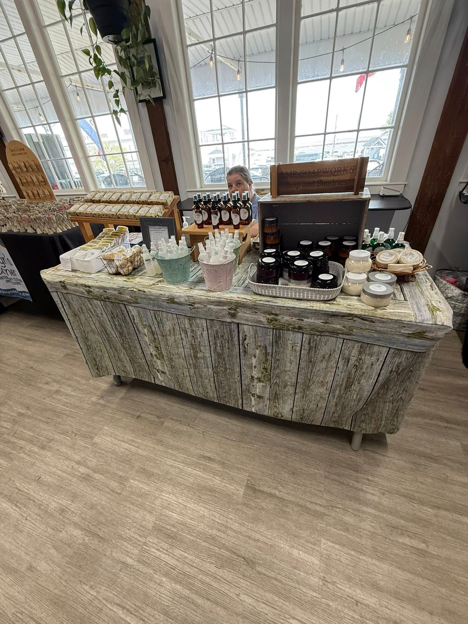 Vendor behind a textured table displaying products. Light-filled room with large windows.