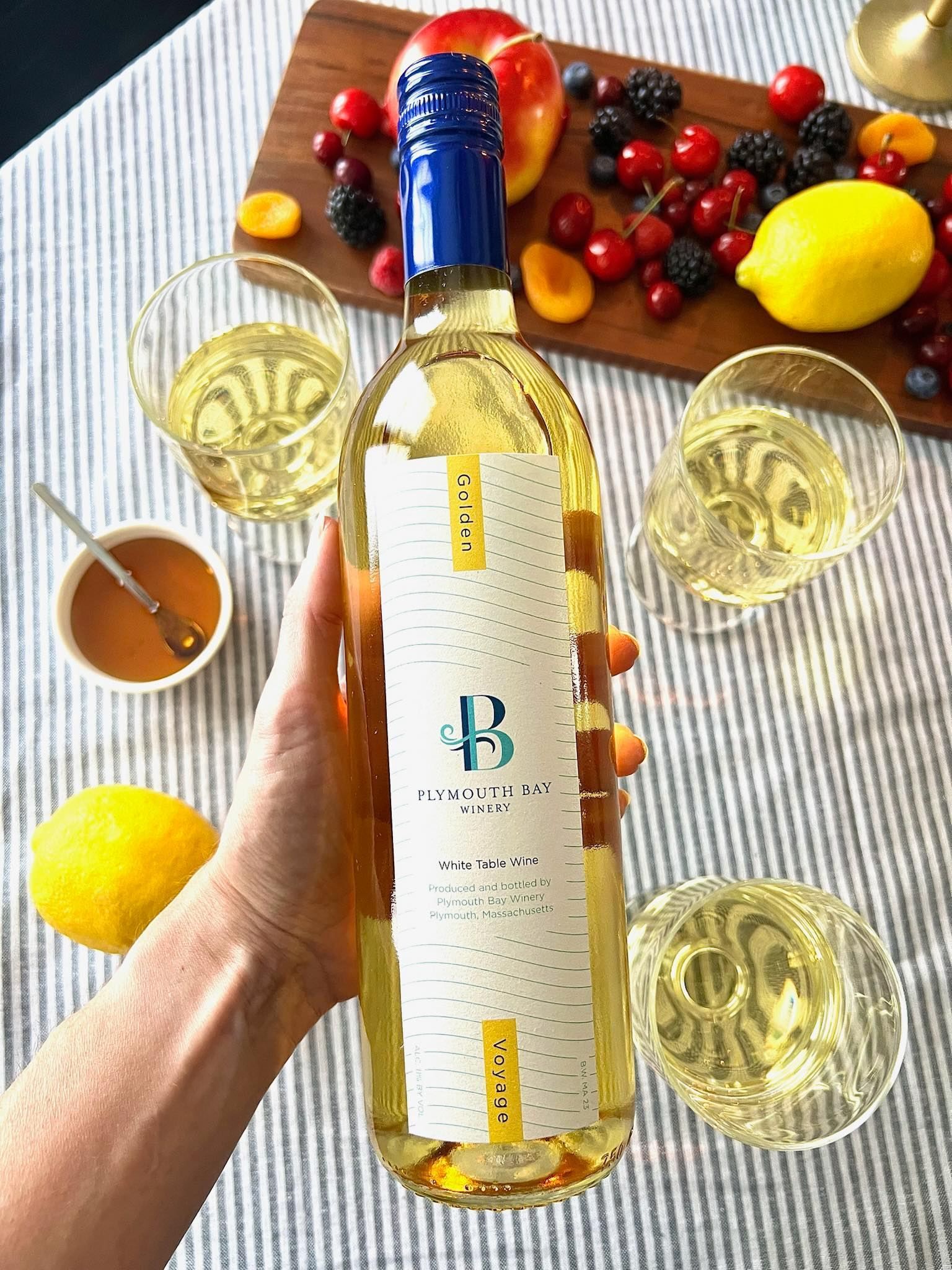 Hand holding white wine bottle with glasses, fruit, and honey on a striped table.