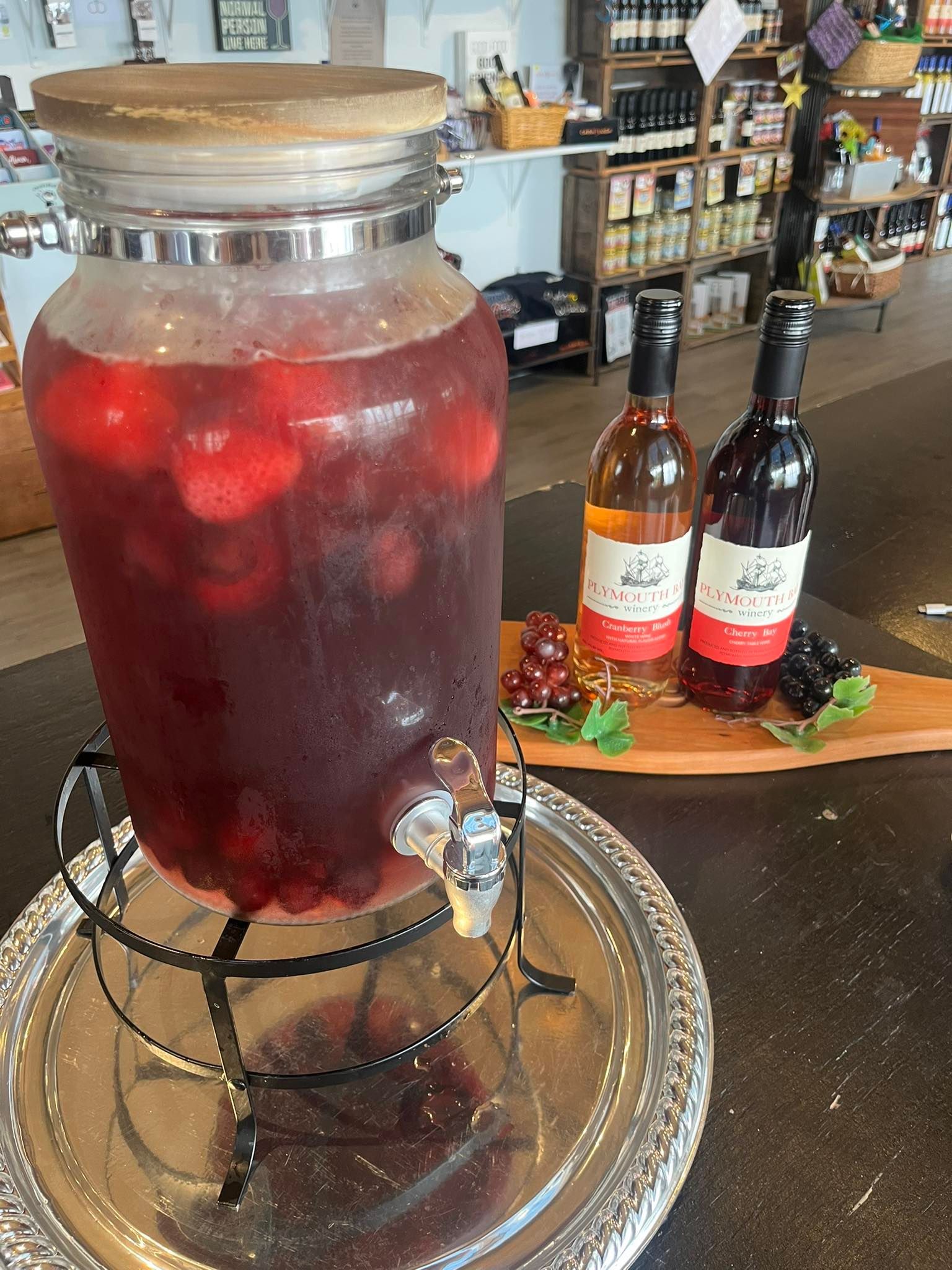 Large beverage dispenser with fruit, two wine bottles, and grapes on a wooden board.