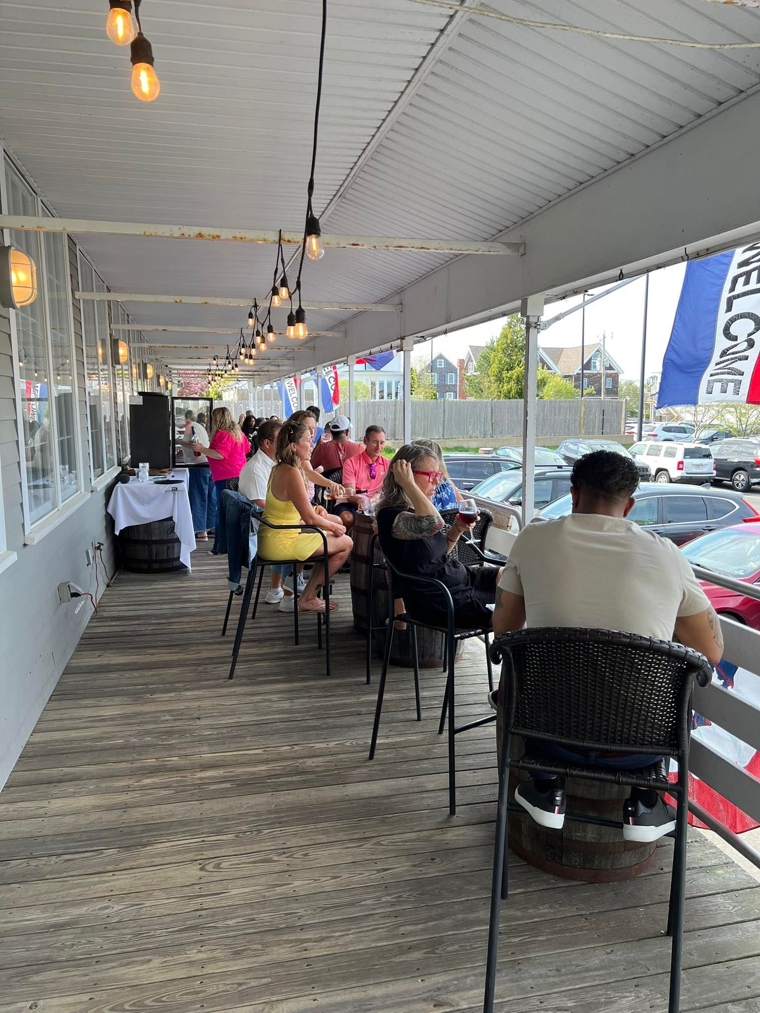 Outdoor restaurant patio with patrons seated at bar stools. Overhead lights and red, white, and blue flags.