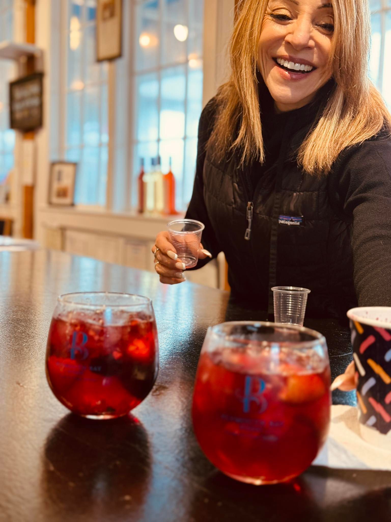 Woman smiles at a table with two red drinks. She holds a small cup and the background is a bright indoor setting.