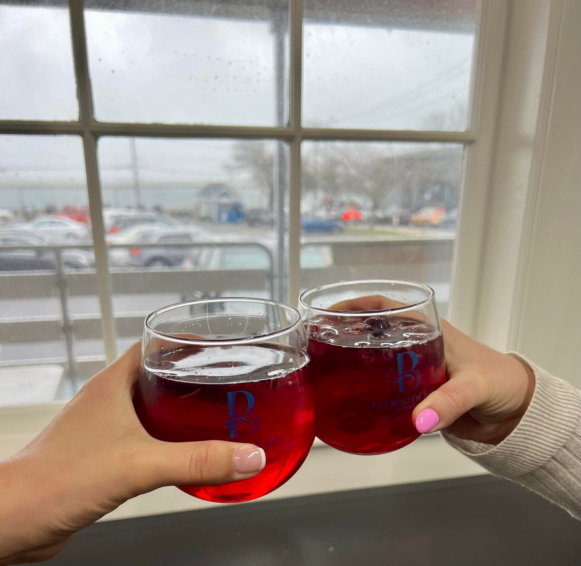 Two stemless wine glasses clinked in a cheers, filled with red liquid, by a window overlooking a parking lot.