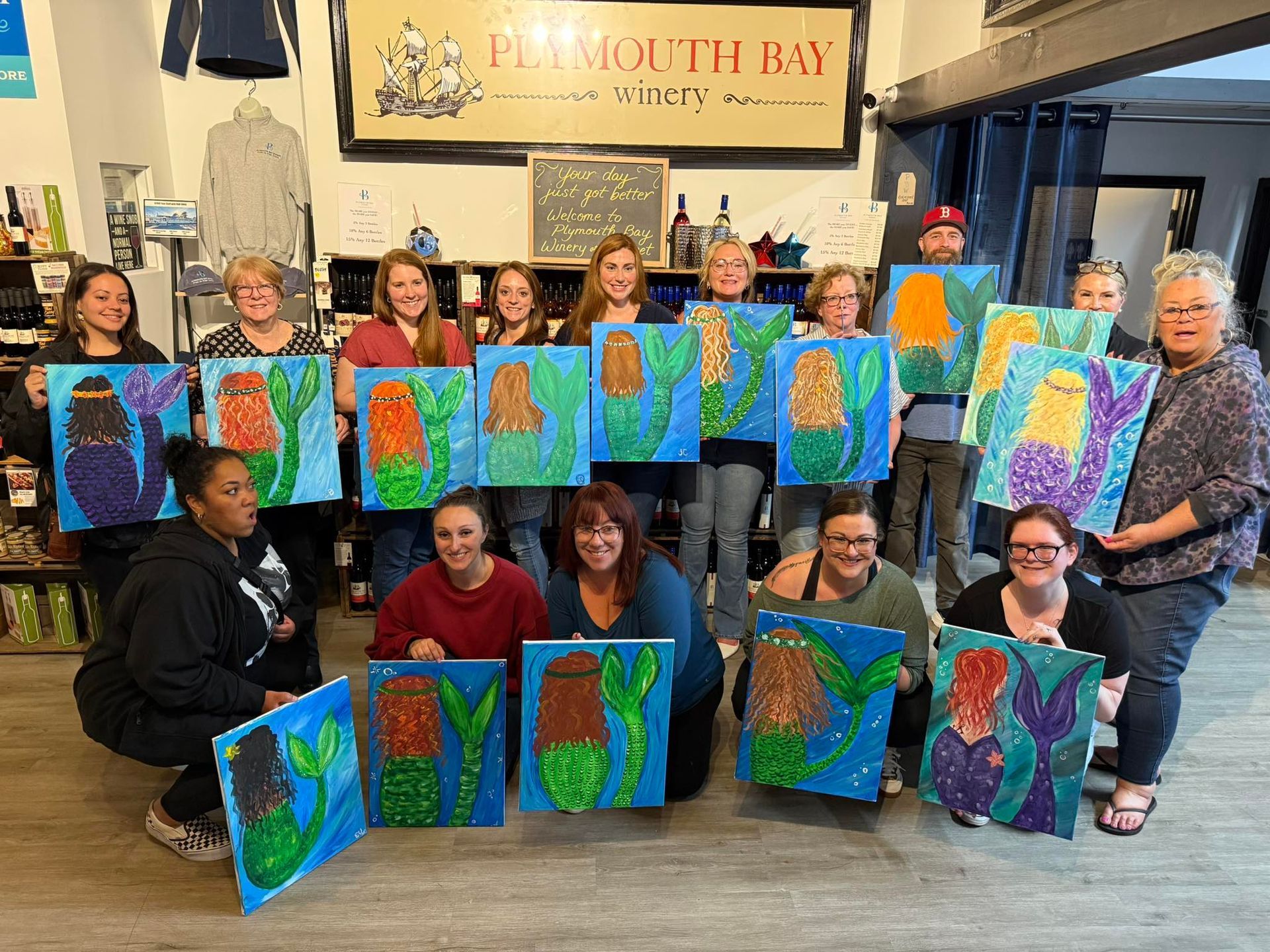 Group of people at Plymouth Bay Winery holding mermaid paintings. Blue water, colorful tails, and green hair.