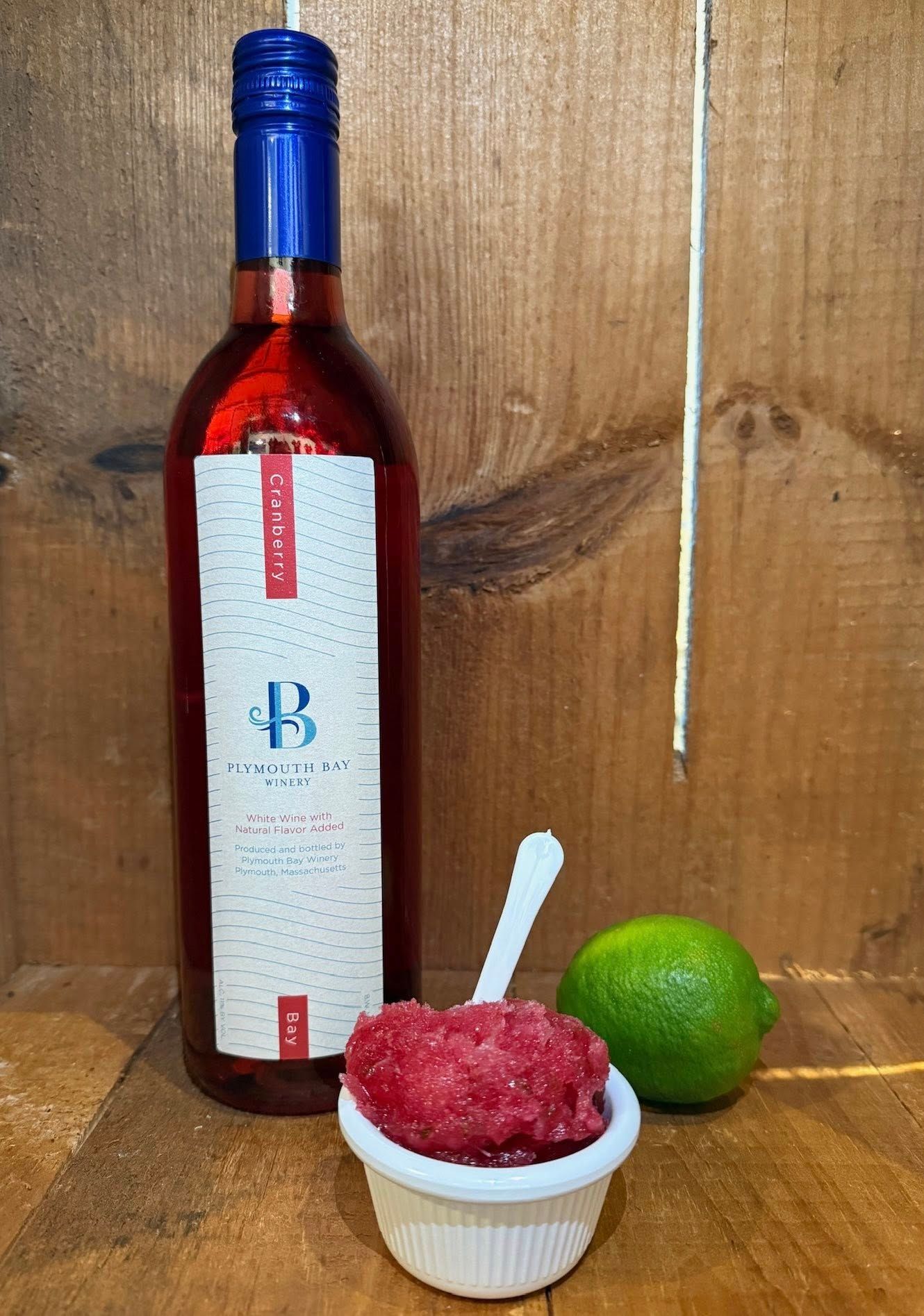 Bottle of red wine, pink slushy in a dish, lime, and spoon on wooden surface.