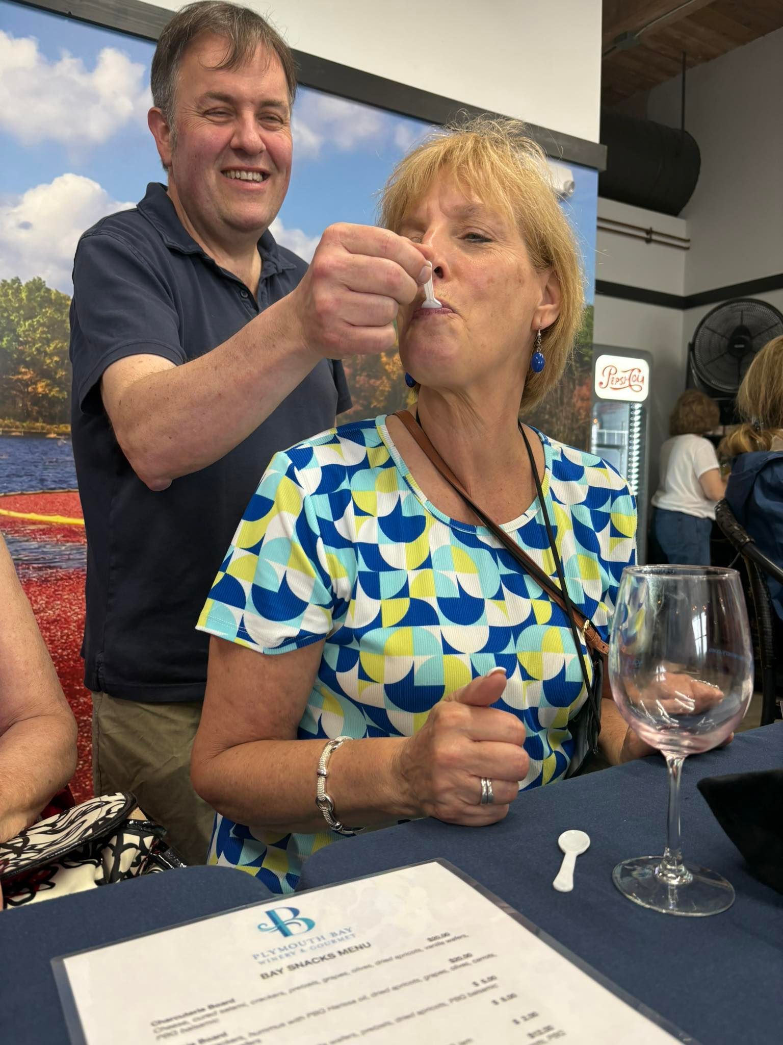 Man offering something on a spoon to a woman. They're at a table with a wine glass, appearing to sample.