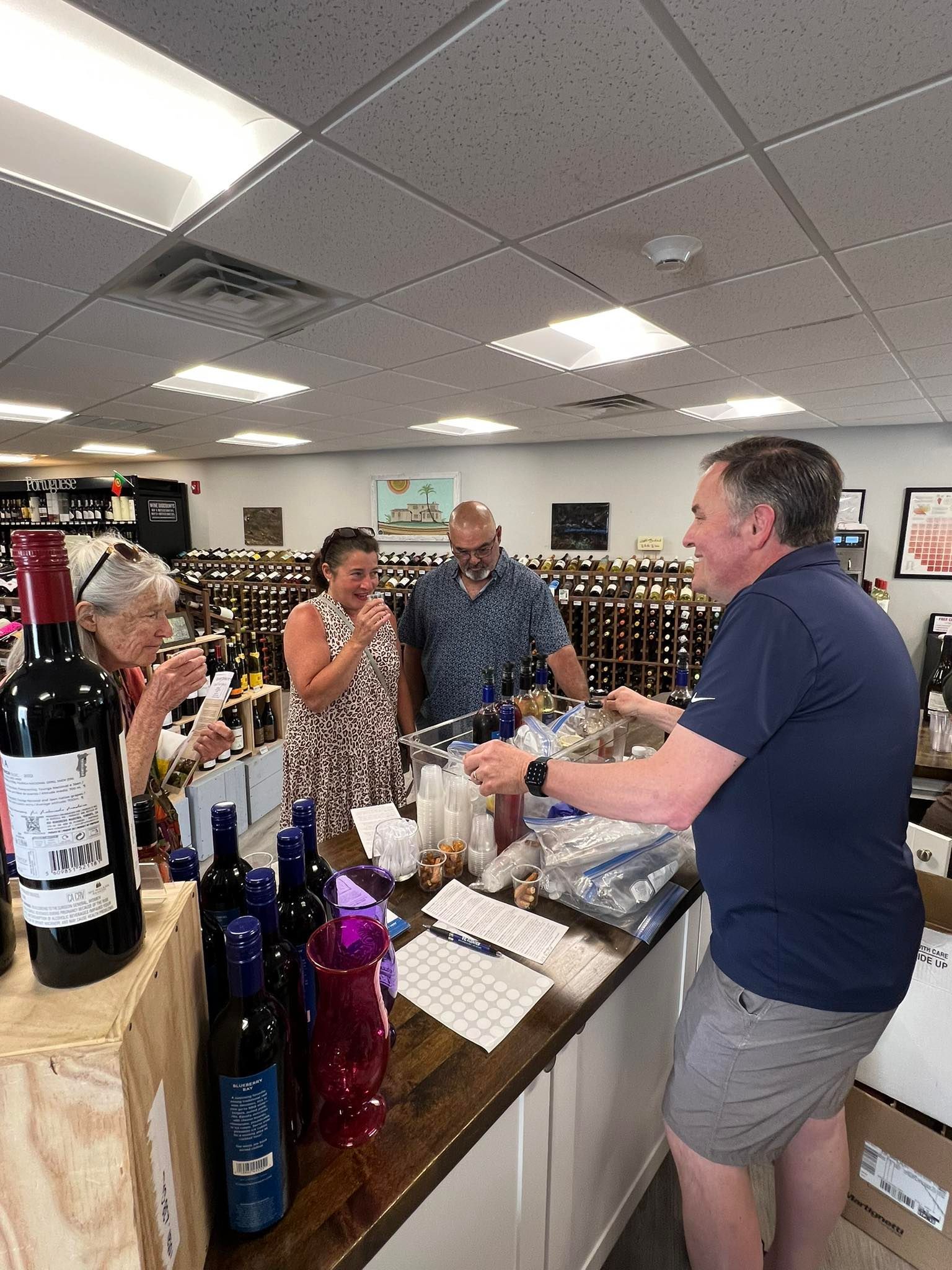 People in a wine shop, looking at bottles with a shop worker. Various bottles and price tags on the counter.