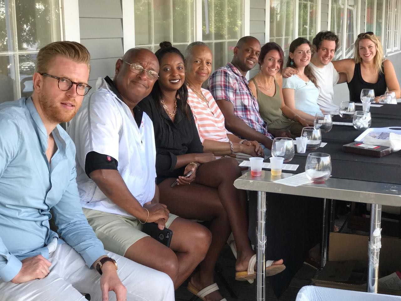 Group of people seated at a table outside, smiling. Glasses and small bowls on table.