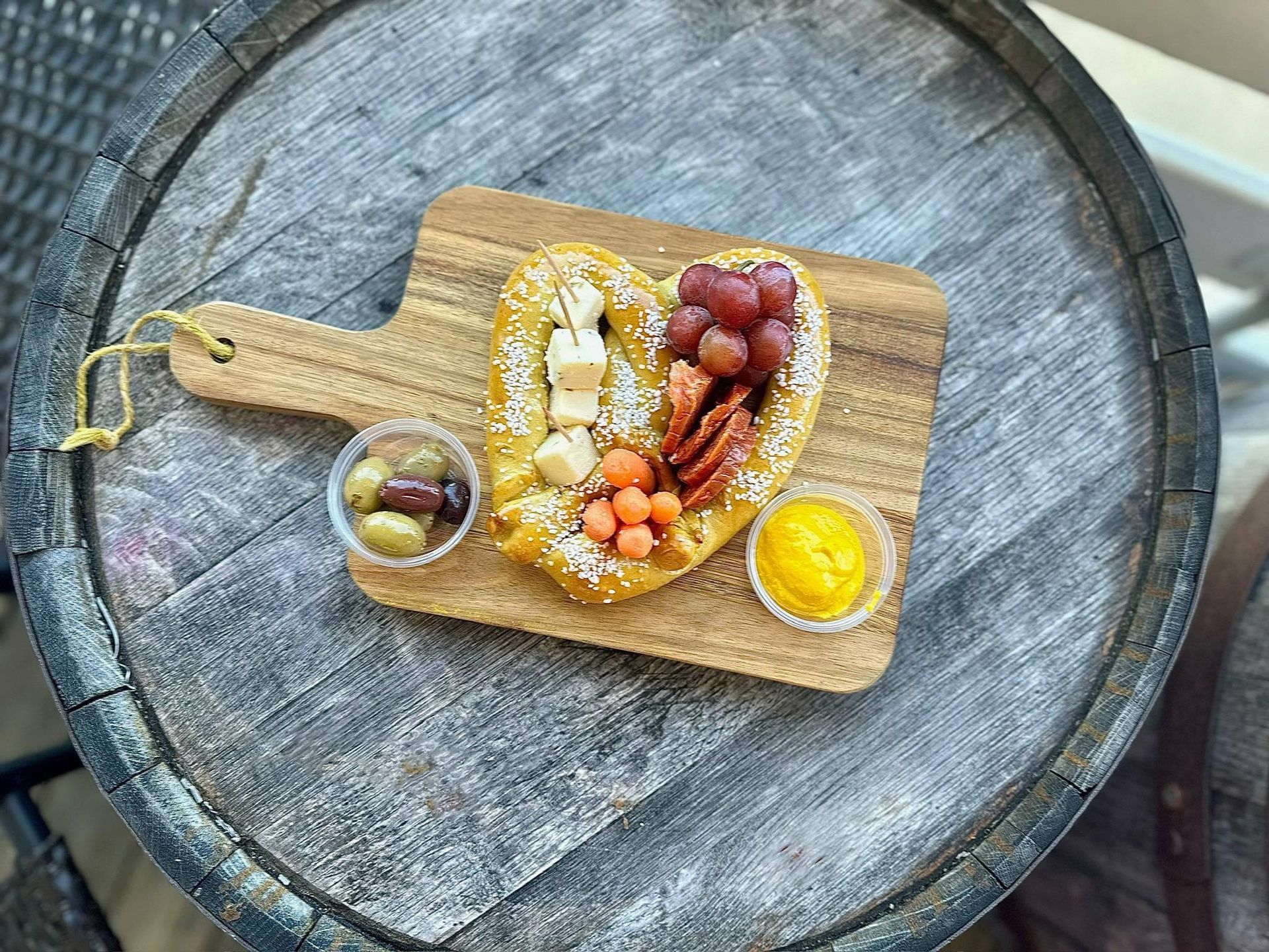 Charcuterie board: heart-shaped pastry, grapes, olives, cheese, mustard, and wood barrel backdrop.