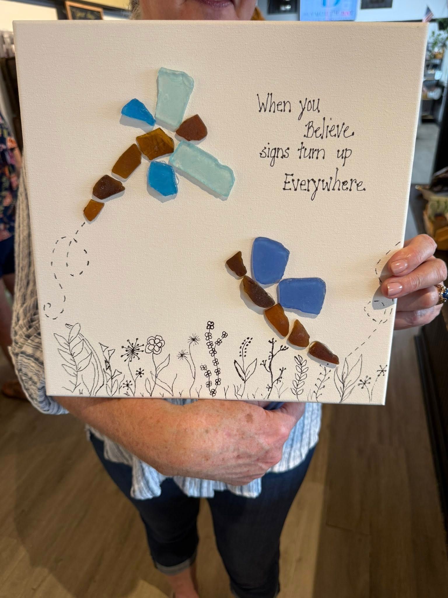 Woman holding a canvas with mosaic dragonflies and text, 