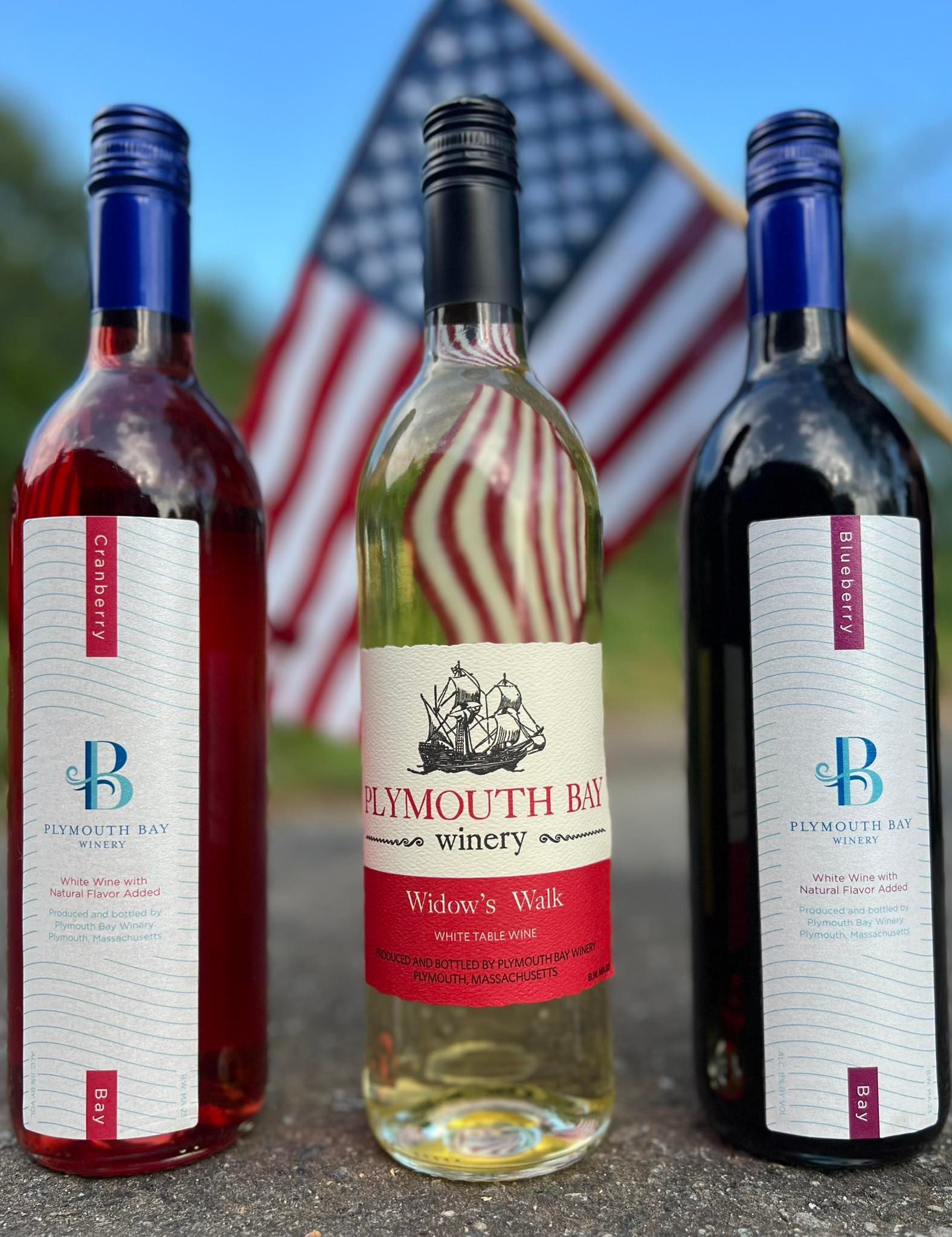 Three wine bottles in front of an American flag; red, white and blue, showcasing the wines from the Plymouth Bay Winery.