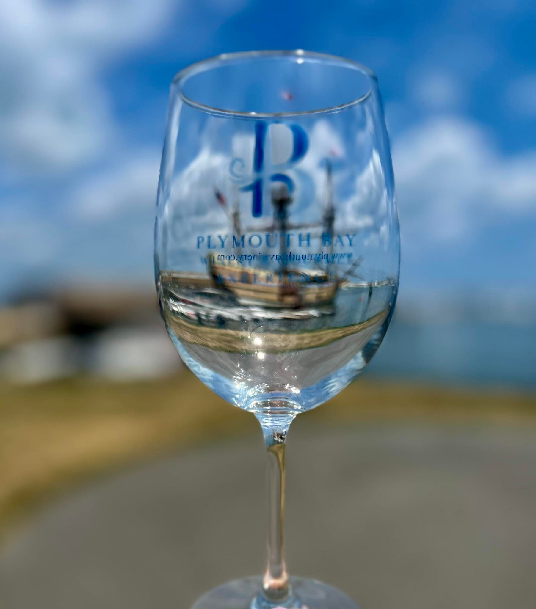 Wine glass reflecting the Mayflower ship at Plymouth Bay, with blue sky and ocean in the background.