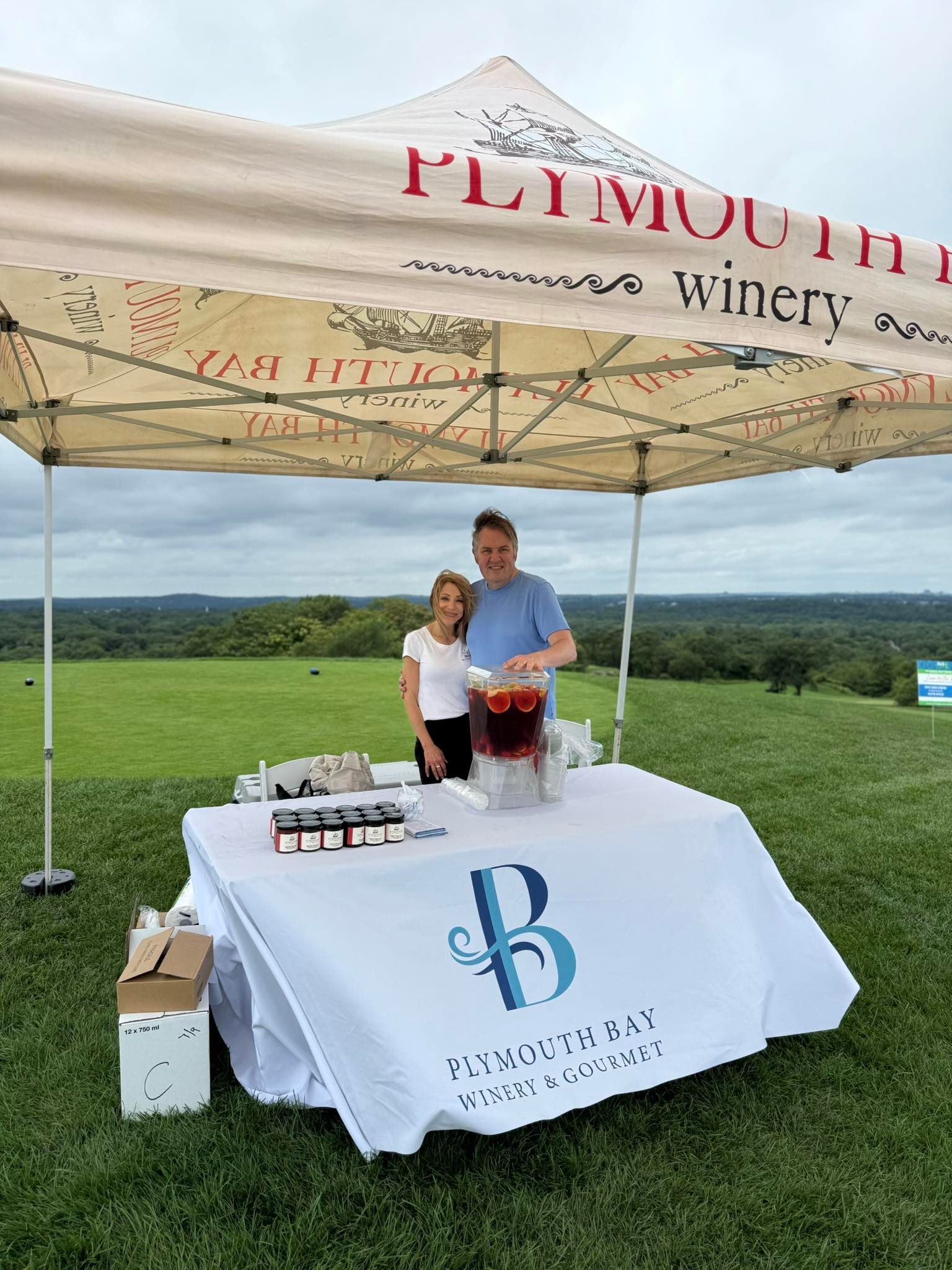 Two people at a Plymouth Winery booth with drinks and a logo on the table, outdoors on a grassy hill.