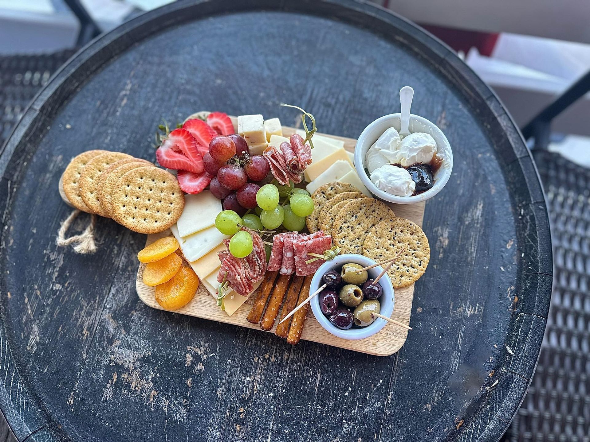 Charcuterie board: crackers, fruit, cheese, olives, and dip on a wooden board atop a dark round table.