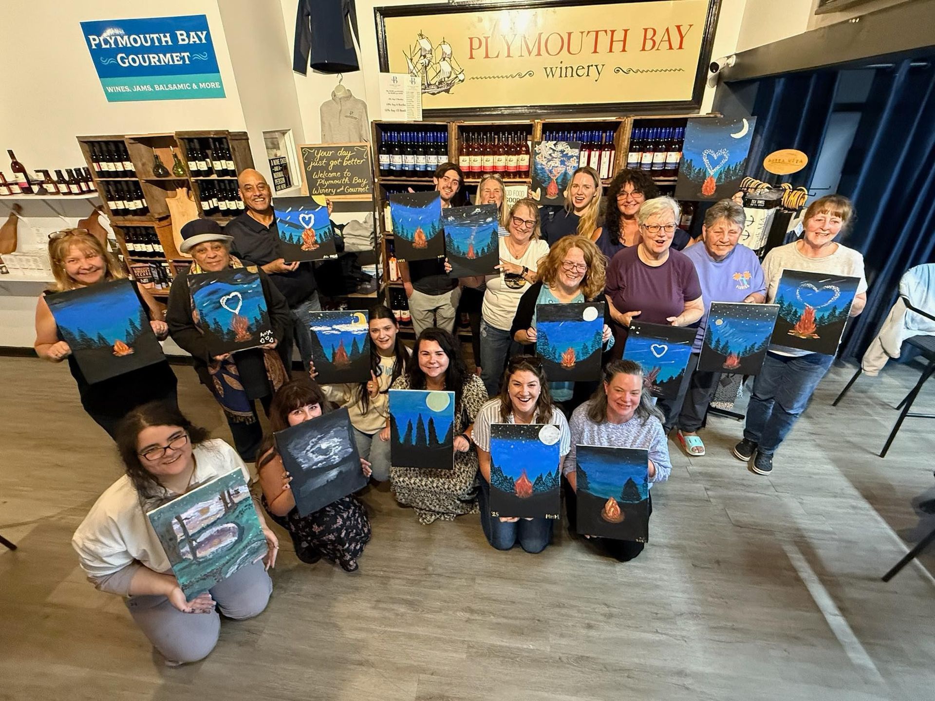 Group of people holding up paintings in a shop. The paintings are dark with a blue sky, trees, and fire.