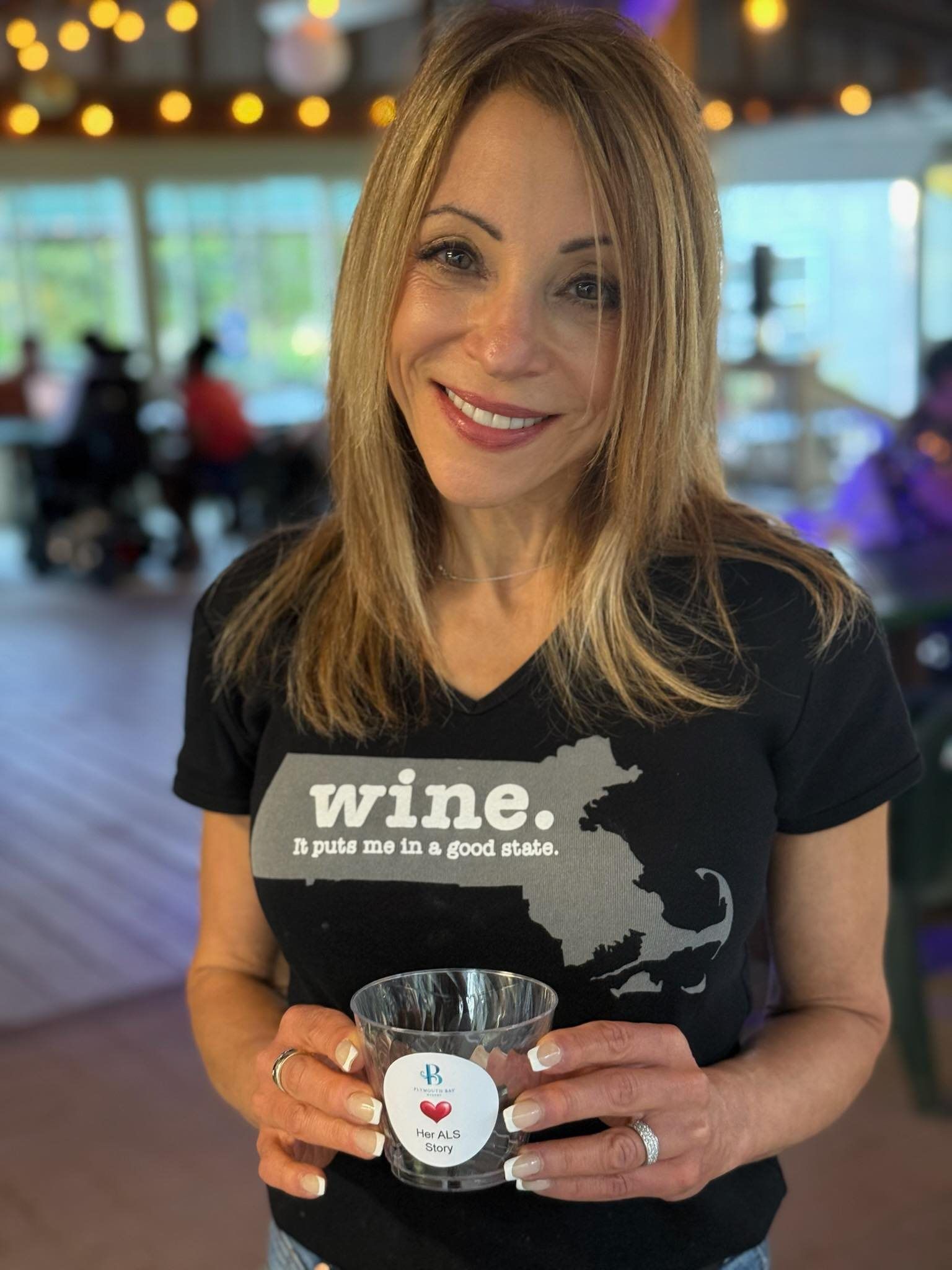 Woman smiling, holding a glass. Wearing a Massachusetts wine shirt, in a barn setting, with string lights and people in the background.