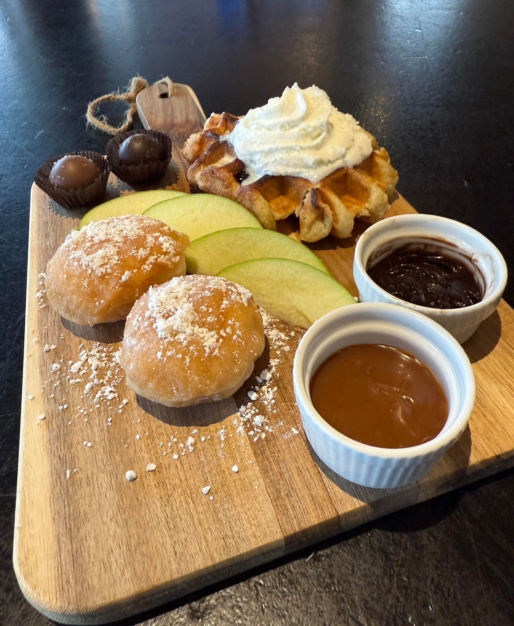 Wooden board with waffle, donuts, apple slices, chocolate, whipped cream, and dipping sauces.