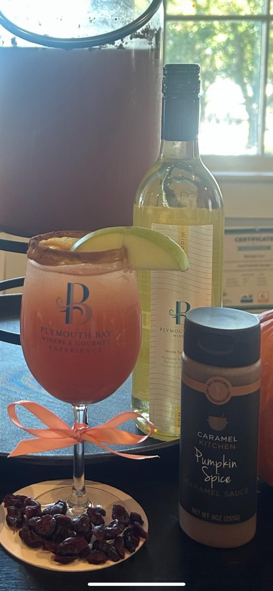 Cocktail with cinnamon rim, apple slice, orange bow, spices, white wine bottle, punch dispenser, on a tray.
