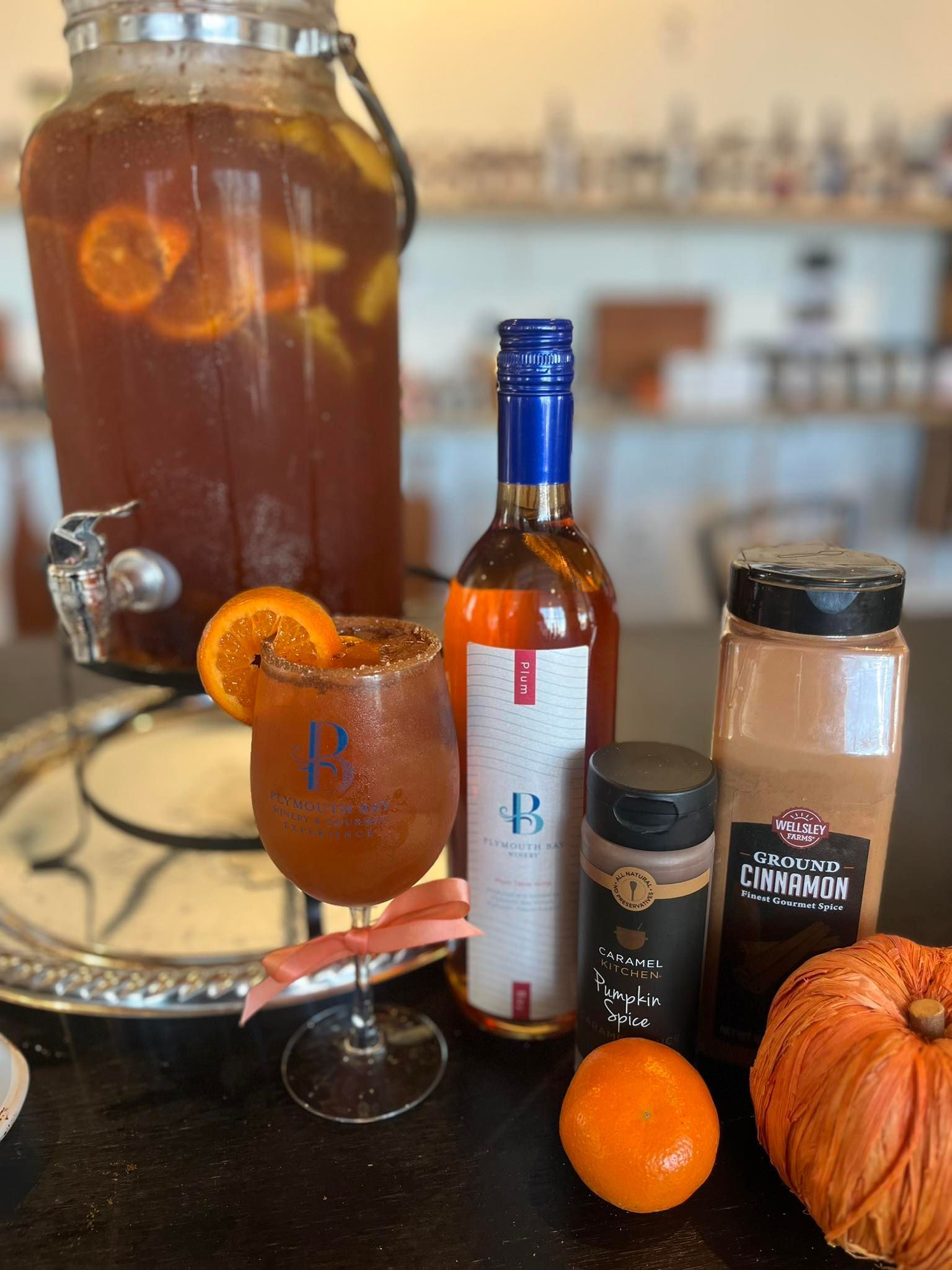 Sangria and ingredients: orange-sliced beverage dispenser, glass with orange slice, wine bottle, spices, and a small pumpkin.