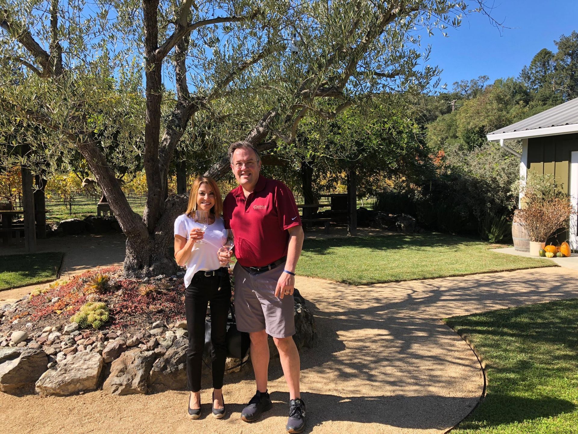 Woman and man holding glasses outdoors near a small building and a stone garden bed.