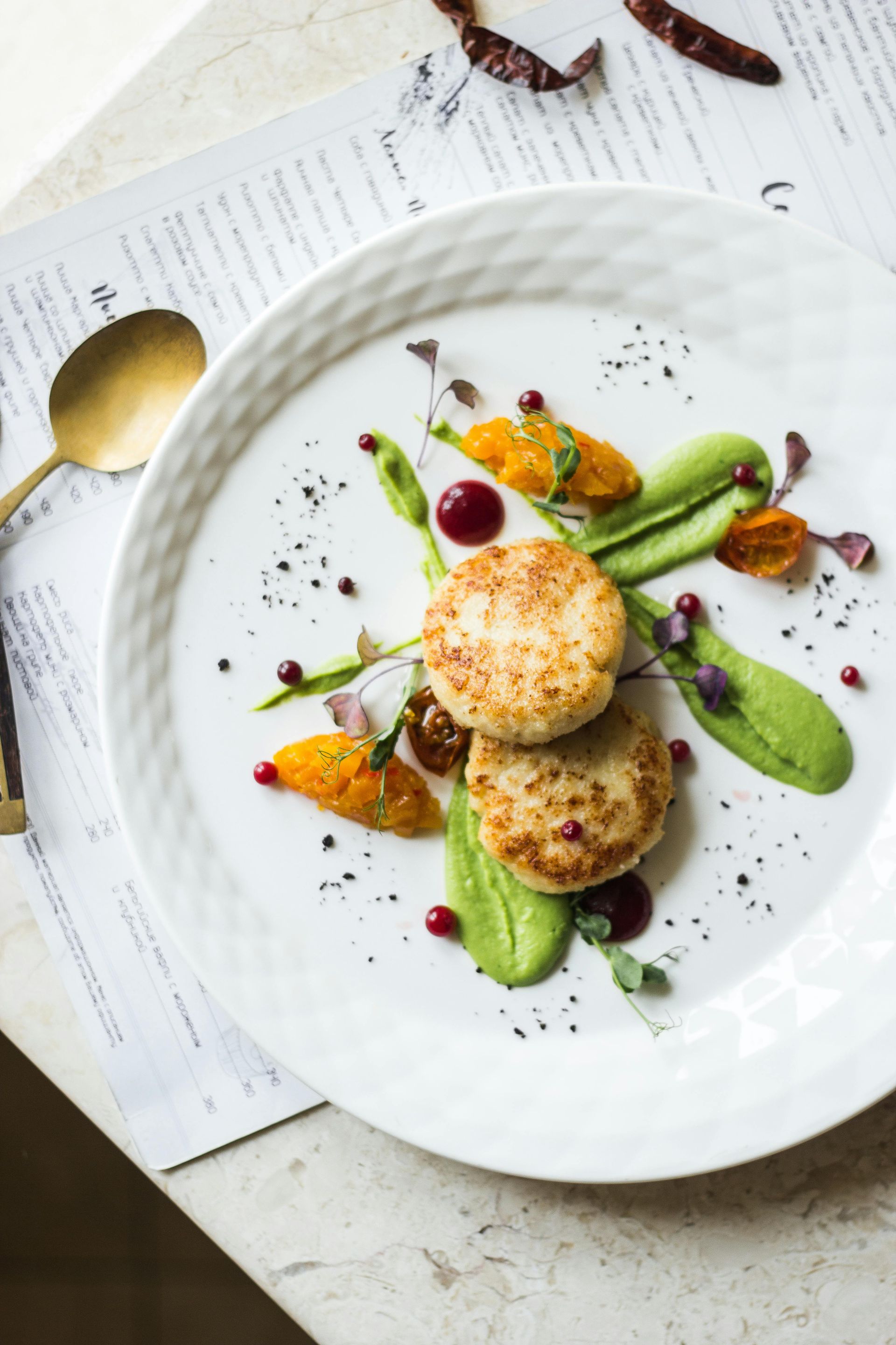 A plate with two crab cakes, pea puree, sweet potato, and garnishes.