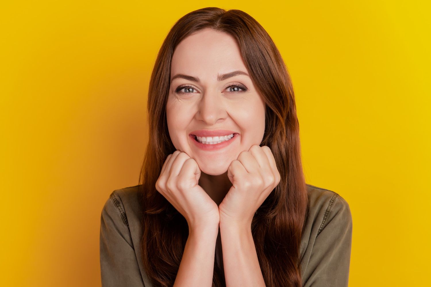 Woman with brown hair smiles, resting her chin on clasped hands, yellow background.