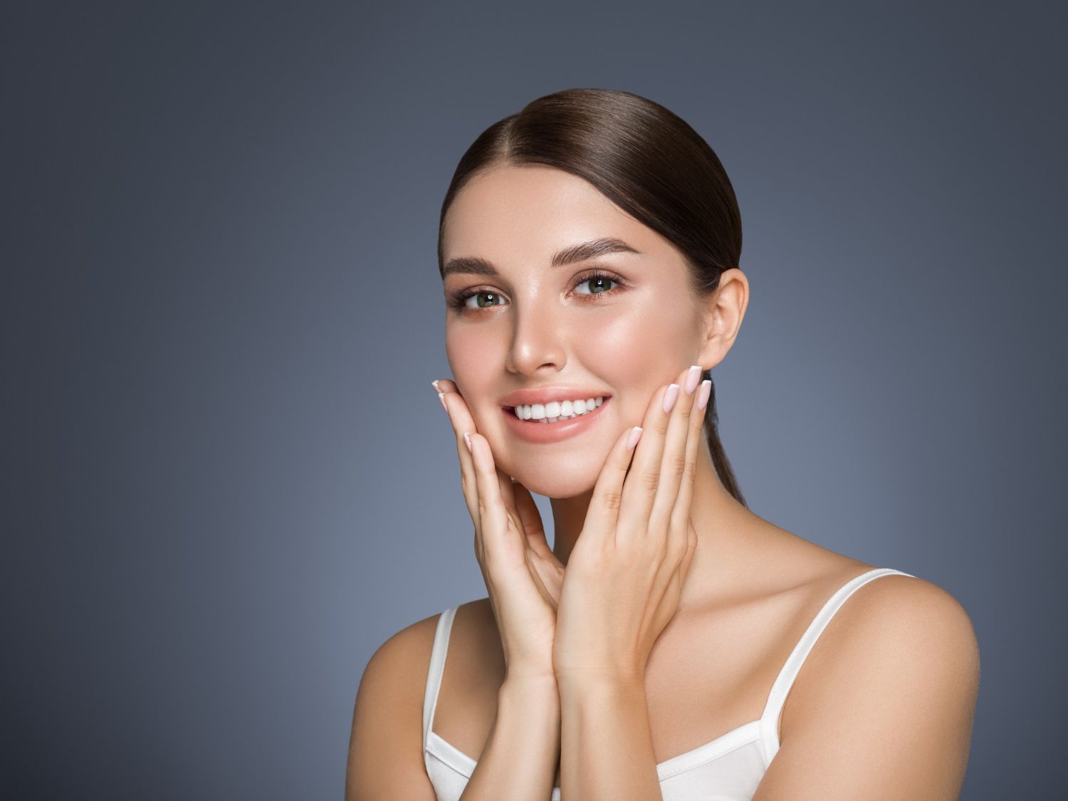 Woman with radiant skin smiling, hands near face, wearing white tank top, against dark blue background.