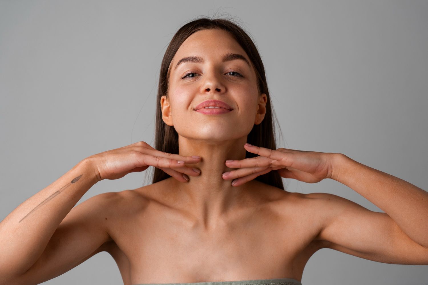 Woman touching her neck, smiling, pale skin, against a gray background.