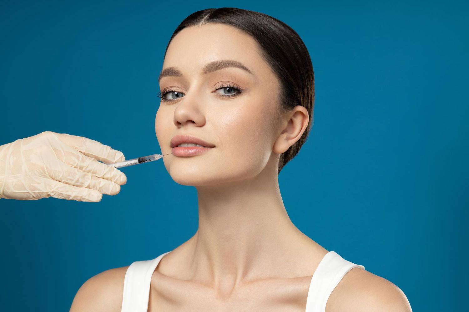 Woman receiving lip injection; a gloved hand holding a syringe. Blue background.
