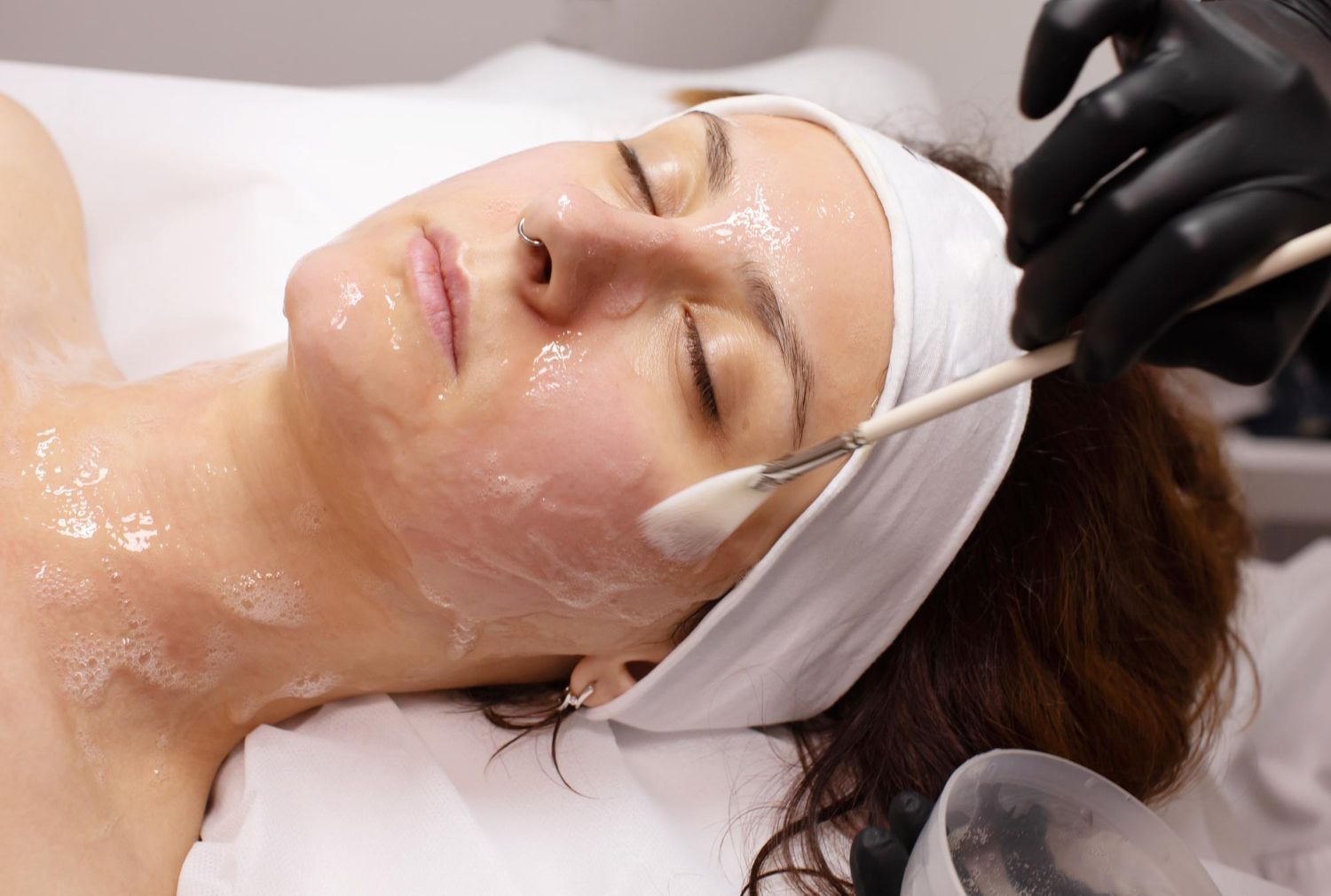 Woman receiving facial treatment with a brush. She wears a headband, eyes closed. Black gloved hand applies product.