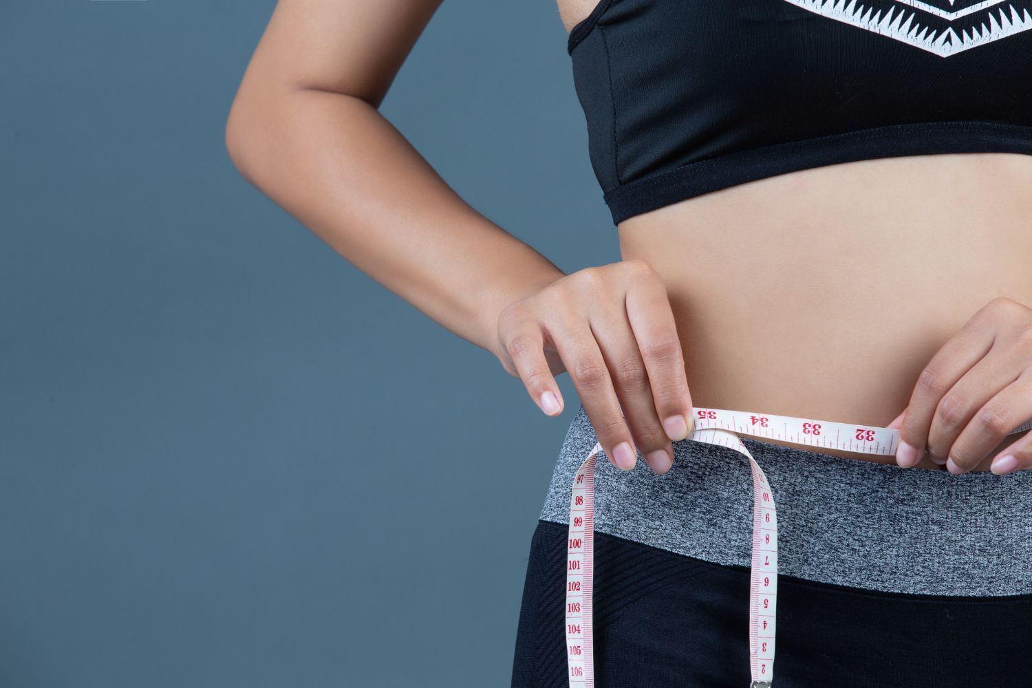 Woman measures her waist with a white and red measuring tape. She is wearing a black sports bra and gray and black leggings.