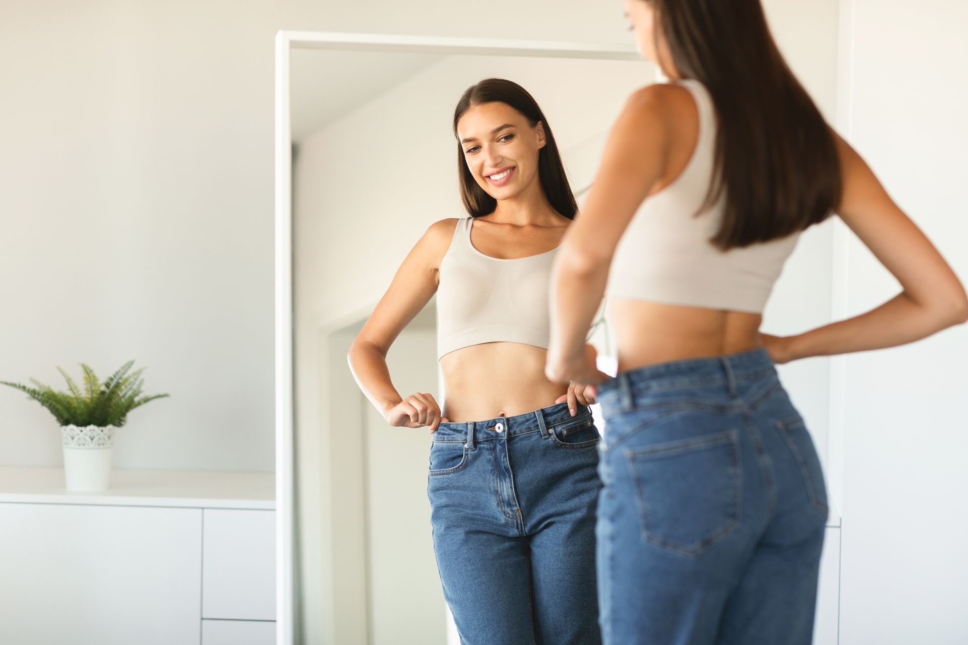 Woman smiles looking at herself in mirror, trying on jeans that are too big.