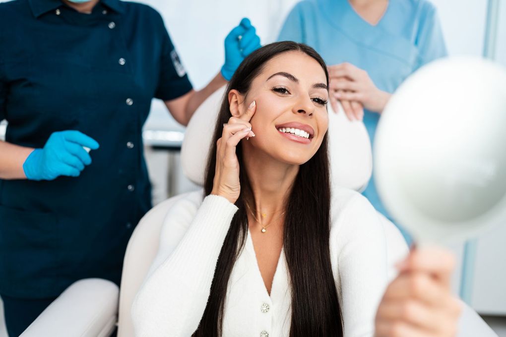 A woman is sitting in a dental chair looking at her face in a mirror.