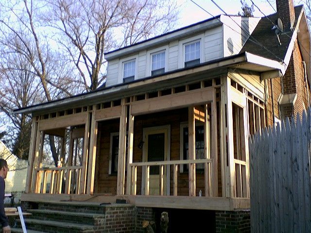 House with a porch under construction. Wooden frame, steps leading up, brick base, and a partially built railing.