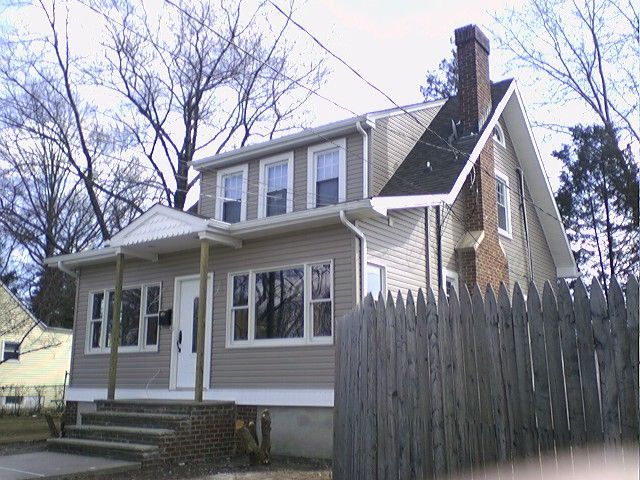 Two-story beige house with a front porch, windows, and a brick chimney, next to a wooden fence.