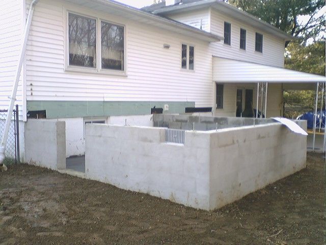 A concrete foundation under construction extends from a white house. Dirt surrounds the structure.