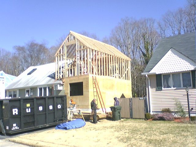 Construction of a two-story building addition; framing stage. Workers, ladder, dumpster, houses. Blue sky.