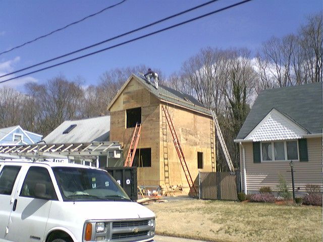 Construction of a two-story building; ladders against the wooden structure, a worker on top.