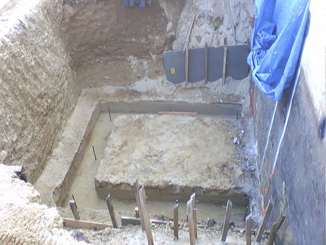 Excavated rectangular pit with concrete foundation being poured; construction site.