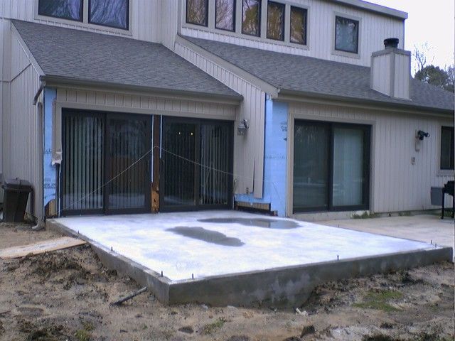 Concrete patio being built outside a light-colored building with sliding glass doors. Dirt and blue insulation visible.