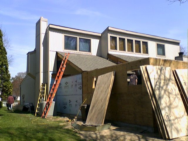 Construction of an addition on a two-story house. Brown plywood, ladders, and a worker are visible.