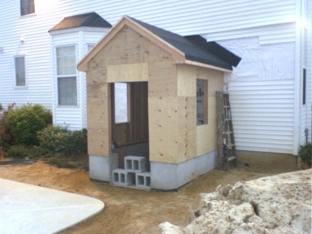 A small unfinished shed with concrete steps, next to a white house. Construction materials present.