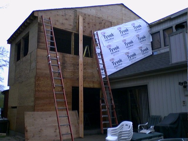 House addition under construction, ladders, plywood siding, Tyvek wrap, outdoor setting.
