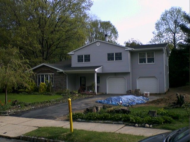 Two-story house with gray siding, two-car garage, and driveway; blue tarp and landscaping materials in front.