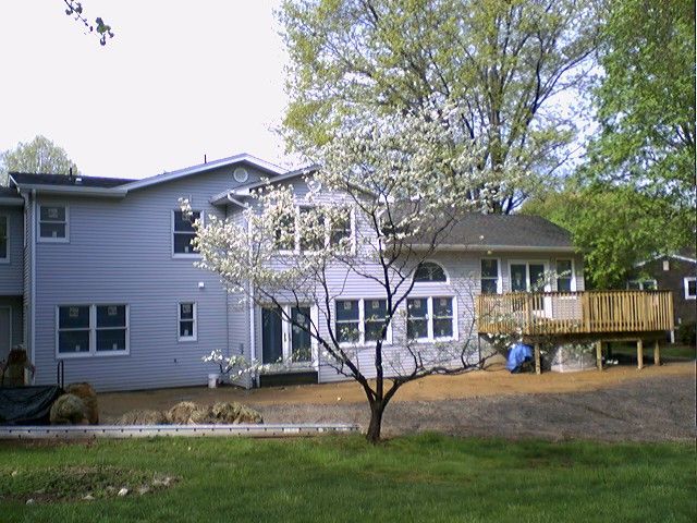 Two-story house with gray siding, white-framed windows, and a wooden deck. A blooming tree is in front.