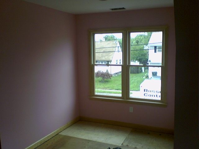 Pink-walled room with window, wooden trim, and unfinished floor, looking out to houses and a green lawn.