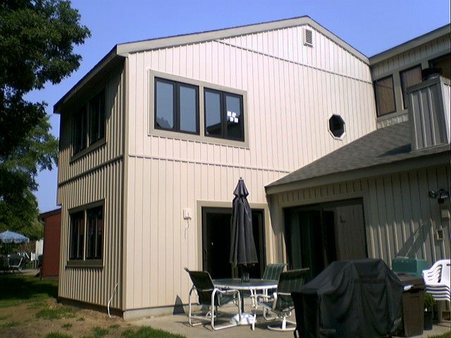 Beige-sided two-story house with dark-framed windows. A patio with outdoor furniture is in front.