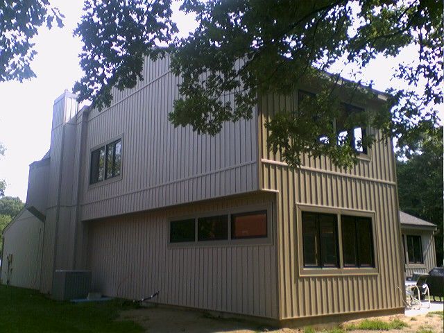Two-story house with vertical siding, windows, and a brick chimney, set in a wooded area.
