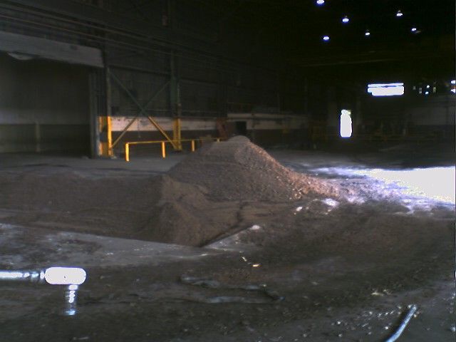 Indoor shot of a large pile of dark material, possibly ore, on a concrete floor inside a factory.